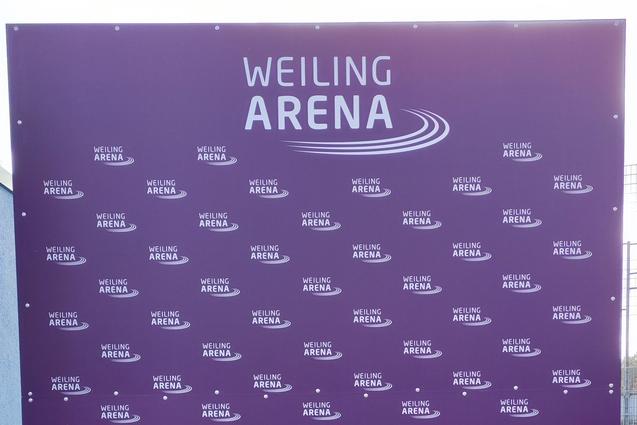 Lila Werbetafel mit wiederholtem Schriftzug "WEILING ARENA" auf grünem Rasen vor einem grauen Gebäude.