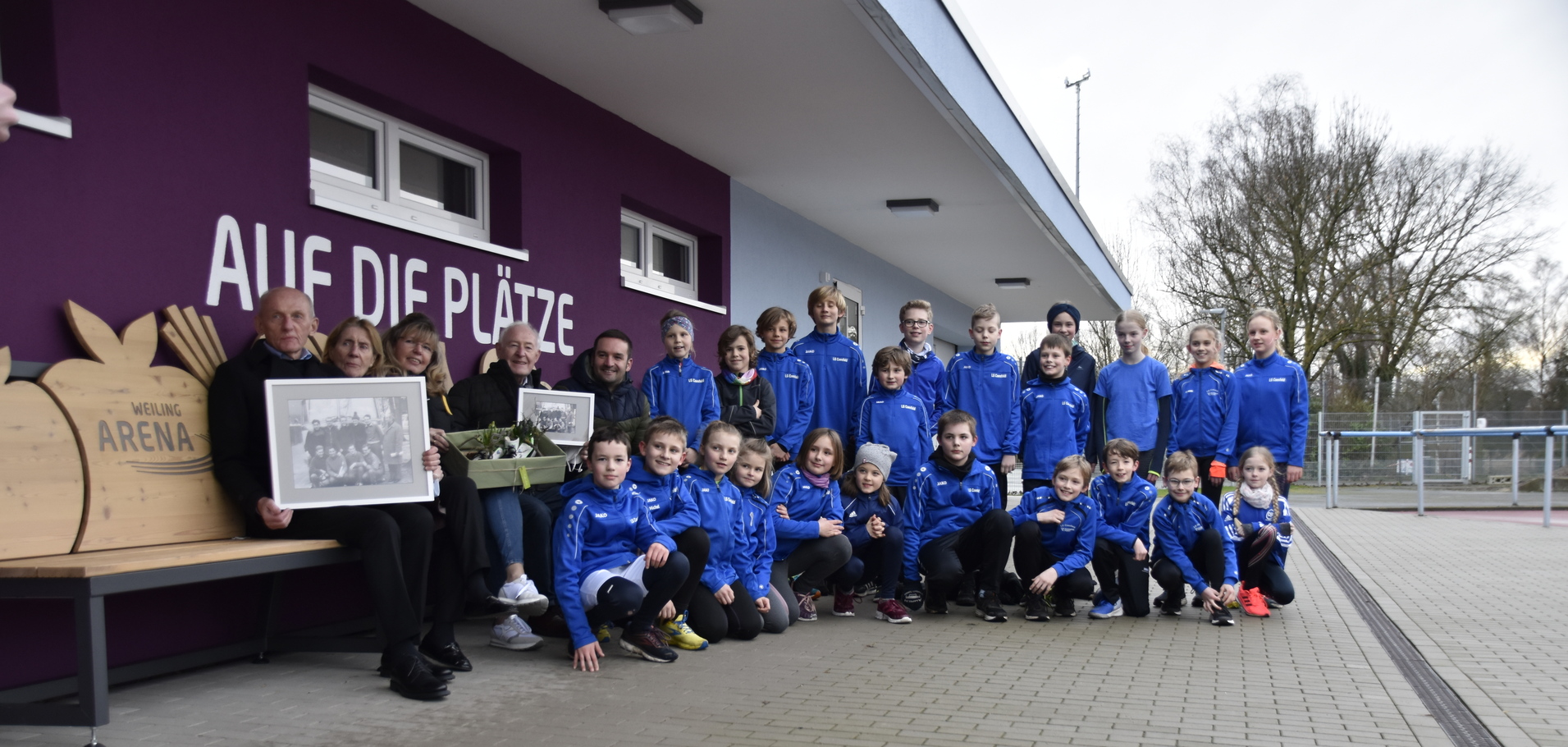 Gruppe von Kindern und Erwachsenen in blauen Sportjacken vor einer Wand mit dem Schriftzug „AUF DIE PLÄTZE“.
