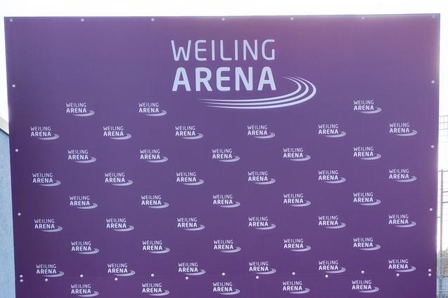 Lila Werbetafel mit wiederholtem Schriftzug "WEILING ARENA" auf grünem Rasen vor einem grauen Gebäude.