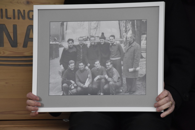 Schwarzweißfoto von neun Männern, die lächeln und posieren, um einen älteren Mann in einem grauen Mantel.