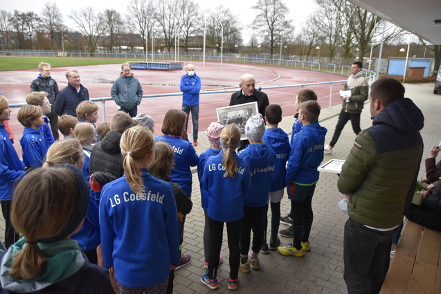 Gruppe von Kindern in blauen Trainingsanzügen hört einem älteren Mann zu, der ein Bild auf einem Sportgelände zeigt.