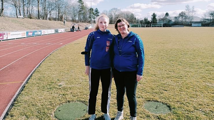 Zwei Frauen in blauen Sportjacken posieren l&auml;chelnd auf einem gr&uuml;nen Fu&szlig;ballfeld mit Laufbahn im Hintergrund.