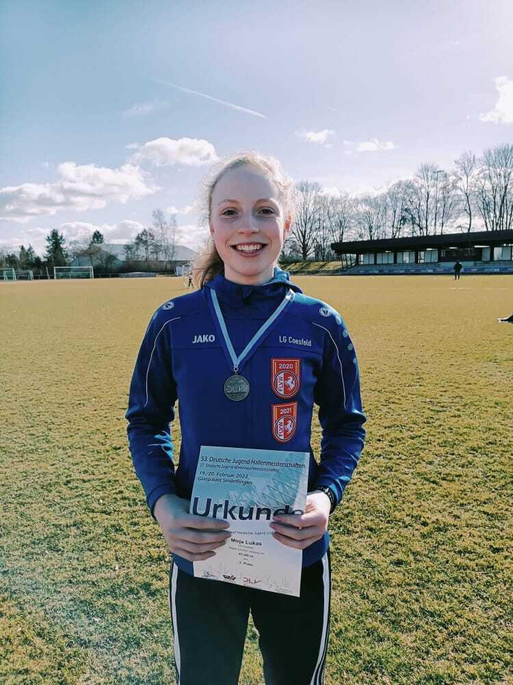 Junge Athletin in blauer Sportjacke hält Urkunde und Medaille auf einem Rasenplatz unter blauem Himmel.