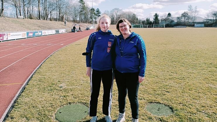 Zwei Frauen in blauen Sportjacken posieren lächelnd auf einem grünen Fußballfeld mit Laufbahn im Hintergrund.