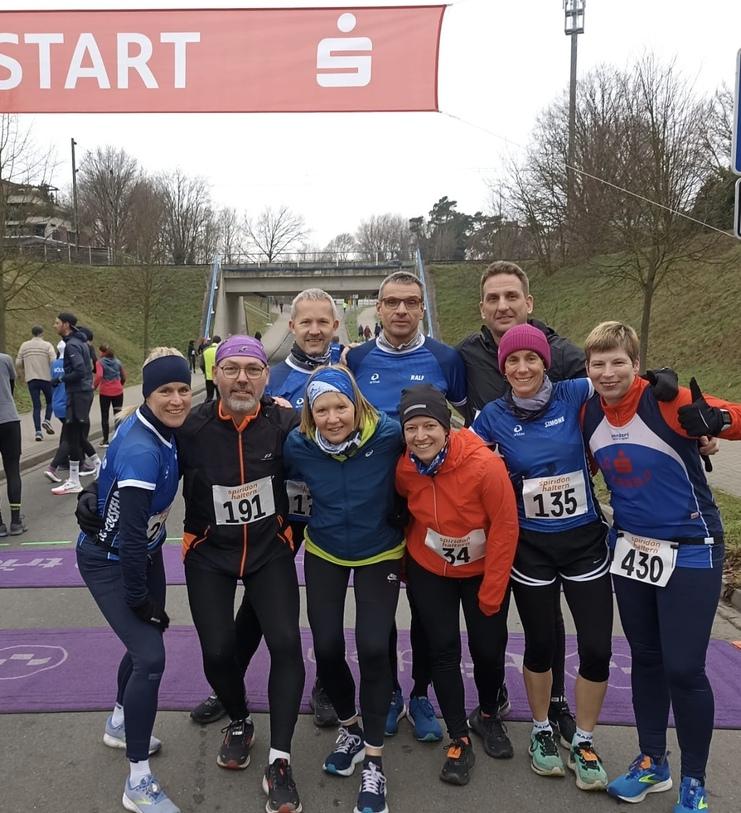 Gruppe von zehn Läufern in sportlicher Kleidung, lächelnd am Start eines Rennens unter einem Banner mit „START“.