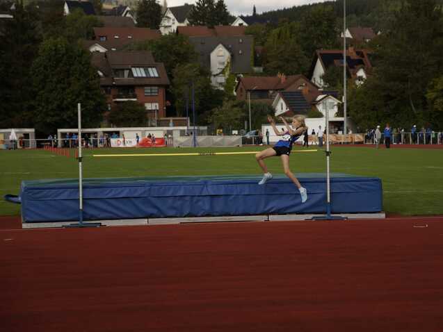 Mädchen springt über eine Hochsprunglatte auf einer Sportanlage, mit Zuschauern und Gebäuden im Hintergrund.