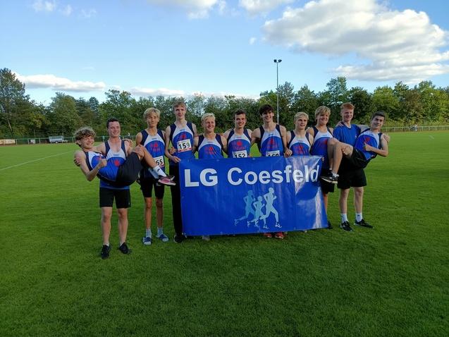Gruppe von Leichtathleten der LG Coesfeld posierend mit einem Banner auf einem Rasenplatz, einer wird angehoben.