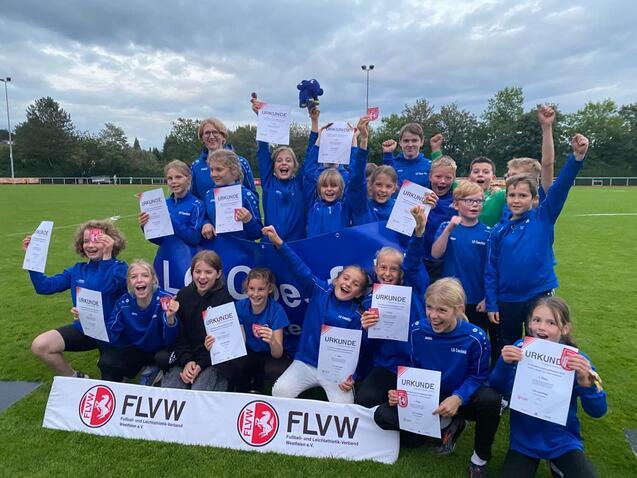 Gruppe von jubilierenden Kindern in blauen Sweatshirts hält Urkunden auf einem Fußballplatz in der Abenddämmerung.