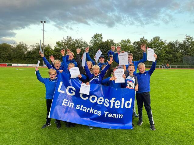Gruppe junger Sportler in blauen Trikots hält Urkunden und ein Banner der LG Coesfeld auf einem Sportplatz hoch.