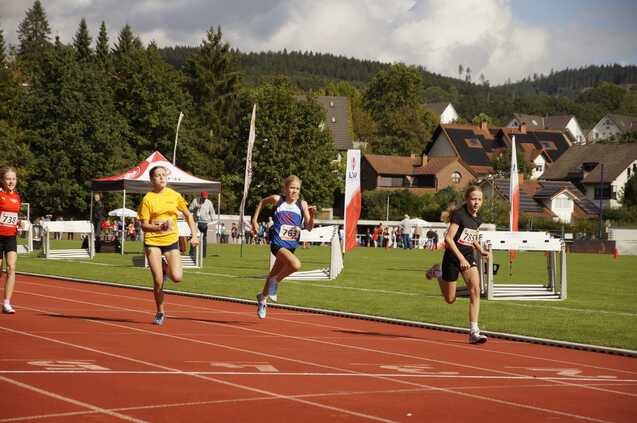 Mädchen in Sportkleidung sprinten auf einer Laufbahn beim Wettkampf, im Hintergrund Zuschauer und Wettkampfanlage sichtbar.