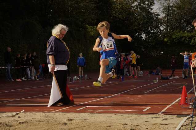 Junge in blauer Sportkleidung springt beim Wettkampf in einen Sandgrube, während ein Schiedsrichter zuschaut.