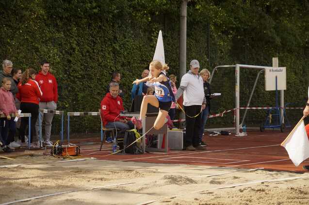 Junge Athletin springt beim Weitsprung über die Sandgrube, Zuschauer und Betreuer beobachten den Wettkampf im Hintergrund.