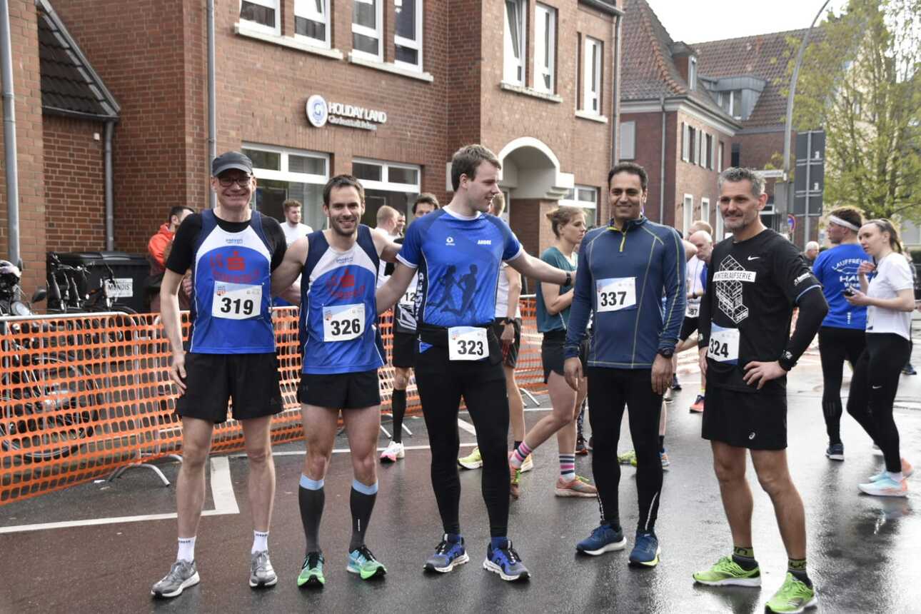 Fünf Läufer in Wettkampfkleidung stehen lässig nebeneinander vor einem Startbereich an einer Straße.