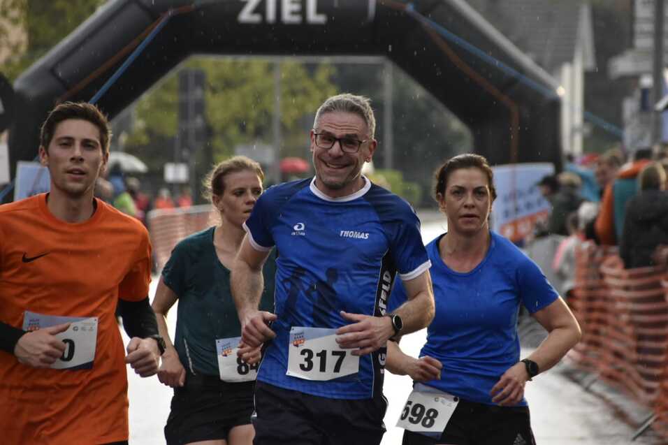 Vier Läufer mit Wettkampfnummern in Sportkleidung laufen unter einem Zielbogen bei regnerischem Wetter.
