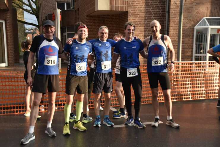 Fünf Läufer in blauen Trikots posieren lächelnd vor einem Wettkampf, umgeben von orangefarbener Absperrung.