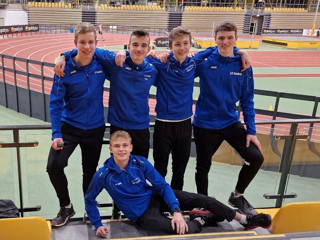 Fünf Jugendliche in blauen Sportjacken posieren lächelnd auf einer Tribüne in einer Sporthalle.