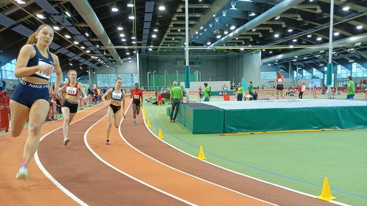 Vier Athletinnen laufen auf einer Hallenbahn, orangefarbene Bahn mit Markierungskegeln, Sporteinrichtungen im Hintergrund.