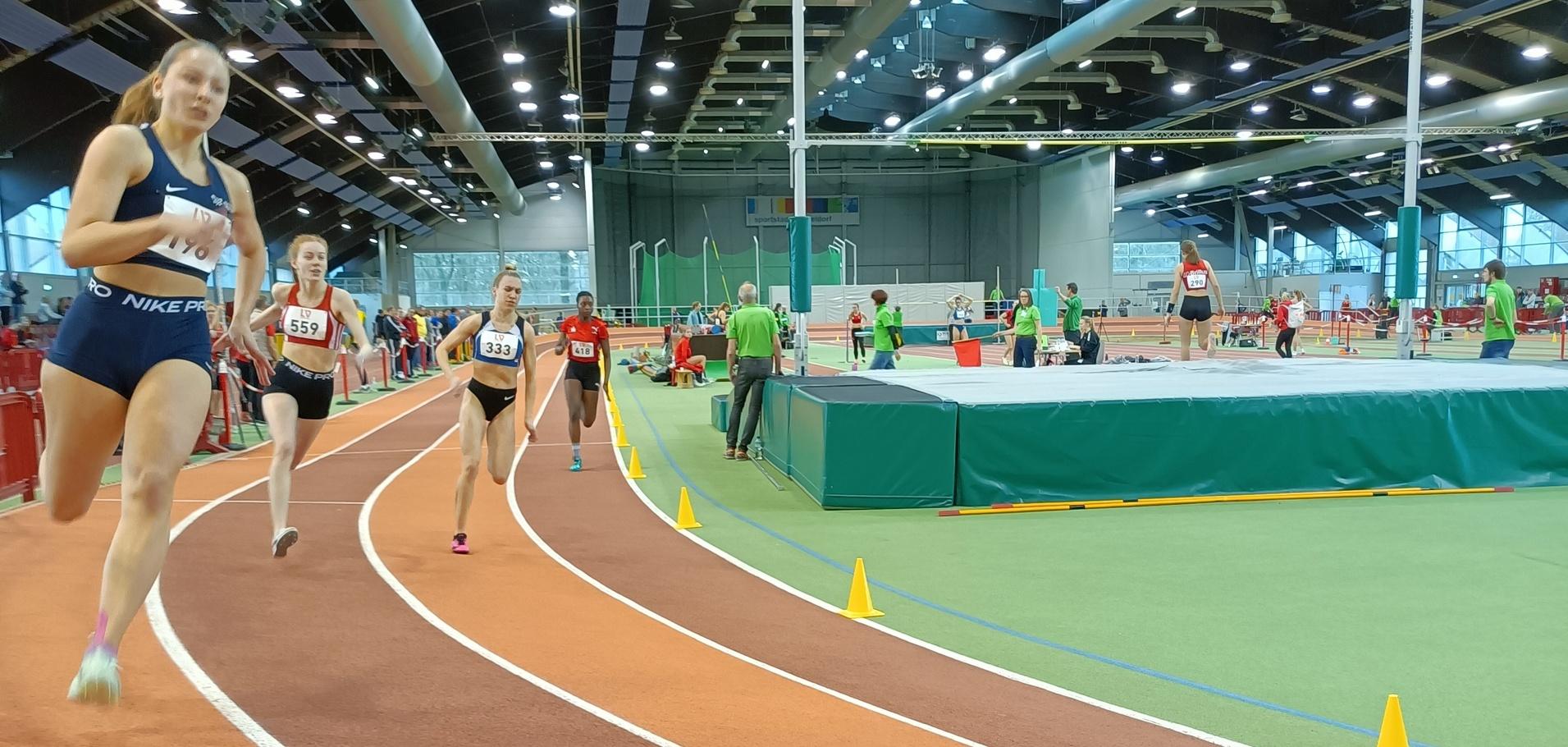 Vier Athletinnen laufen auf einer Hallenbahn, orangefarbene Bahn mit Markierungskegeln, Sporteinrichtungen im Hintergrund.