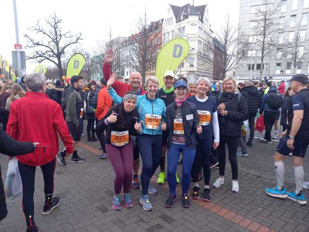 Gruppe von Läufern in Sportkleidung mit Zahlennummern vor dem Start eines Laufereignisses, umgeben von Zuschauern.