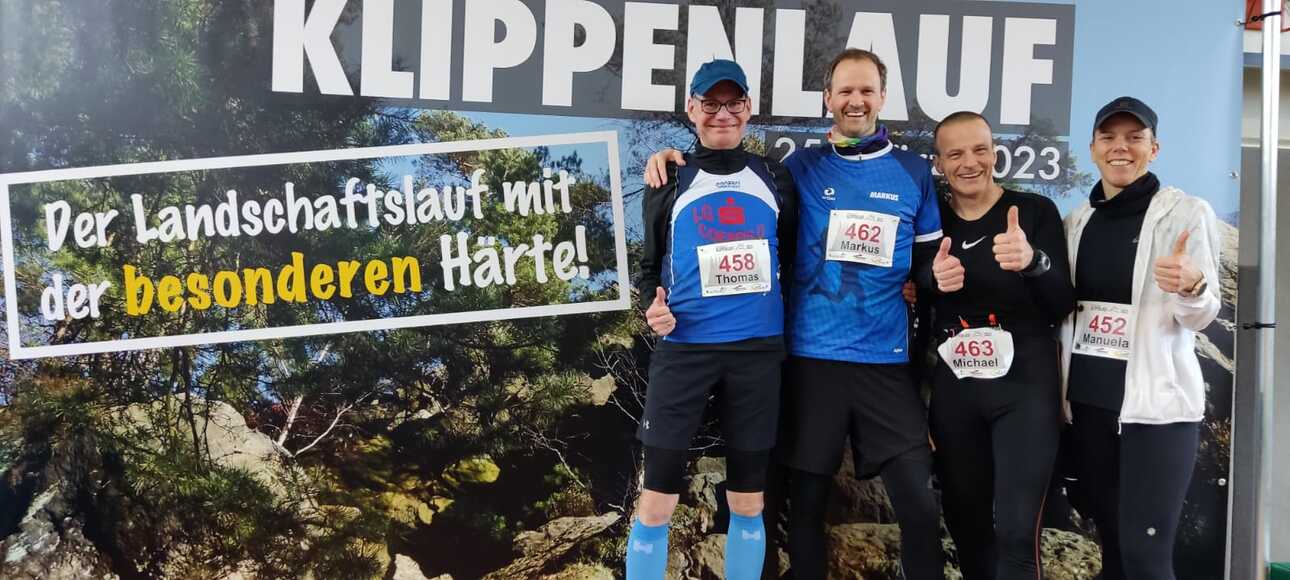 Vier Teilnehmer stehen mit Daumen hoch vor einem Plakat des Klippenlaufs, das eine Landschaft mit Bergen zeigt.