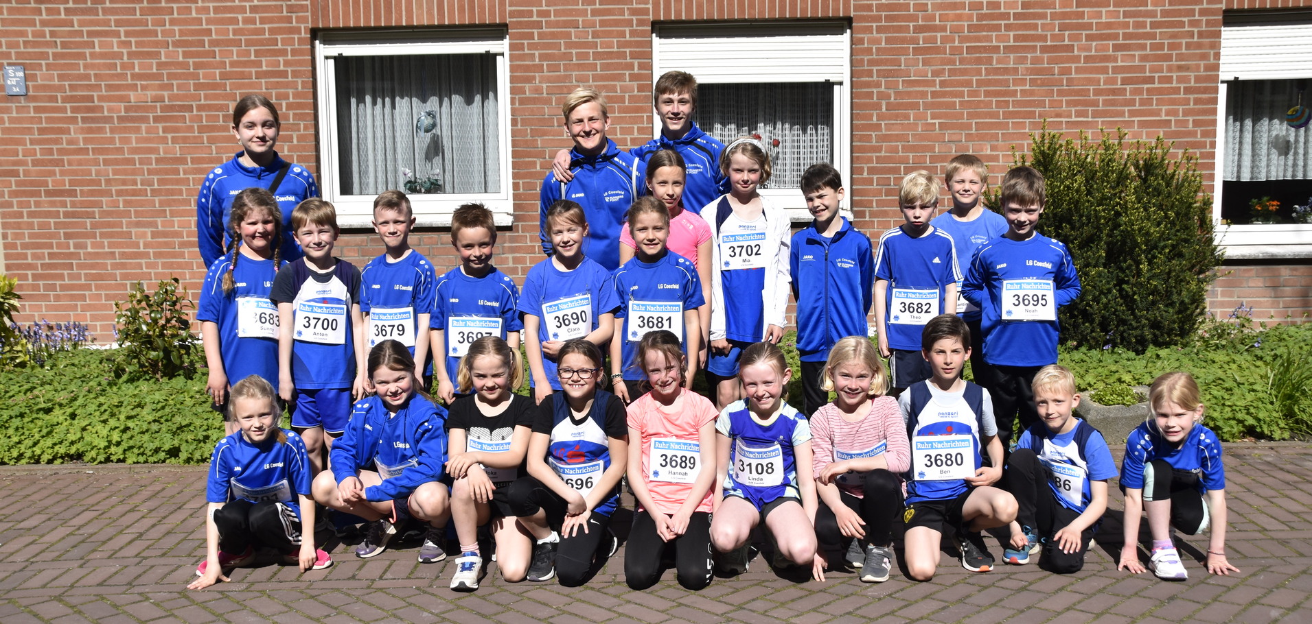 Gruppenfoto von Kindern in blauen Trikots mit Startnummern vor einem Ziegelgebäude, umgeben von grünen Pflanzen.