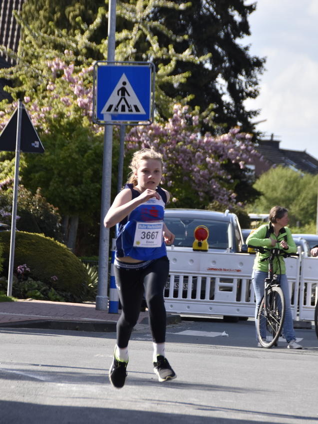 Zwei Kinder laufen in Sportkleidung während eines Wettlaufs auf einer Straße, Zuschauer und Fahrräder im Hintergrund.