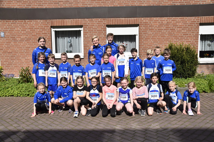 Gruppenfoto von Kindern in blauen Trikots mit Startnummern vor einem Ziegelgebäude, umgeben von grünen Pflanzen.