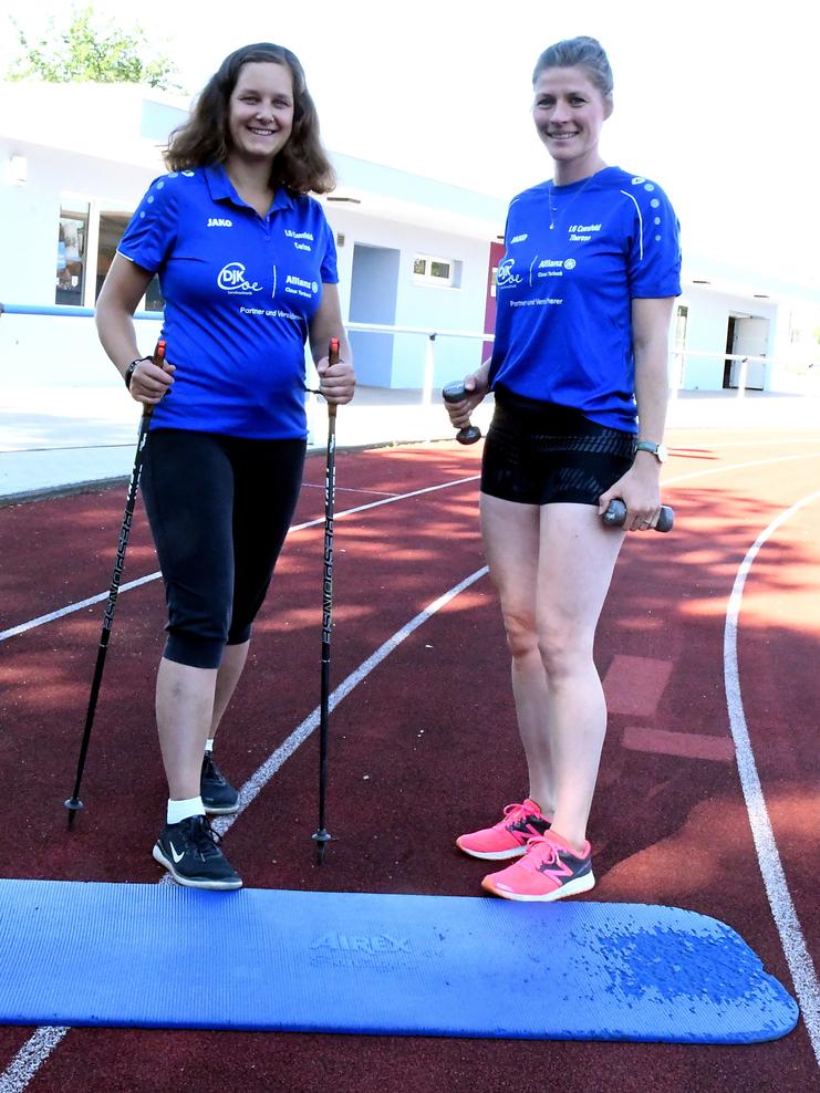 Zwei Frauen in Sportkleidung stehen auf einer Laufbahn, eine mit Nordic-Walking-Stöcken, die andere mit Hanteln.