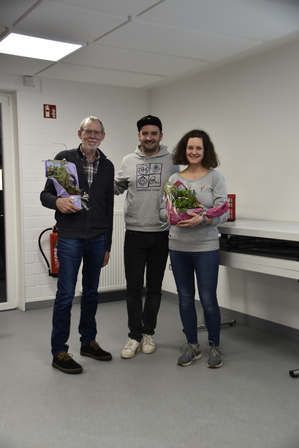 Drei Personen stehen in einem Raum und halten topfblühende Pflanzen in den Händen.