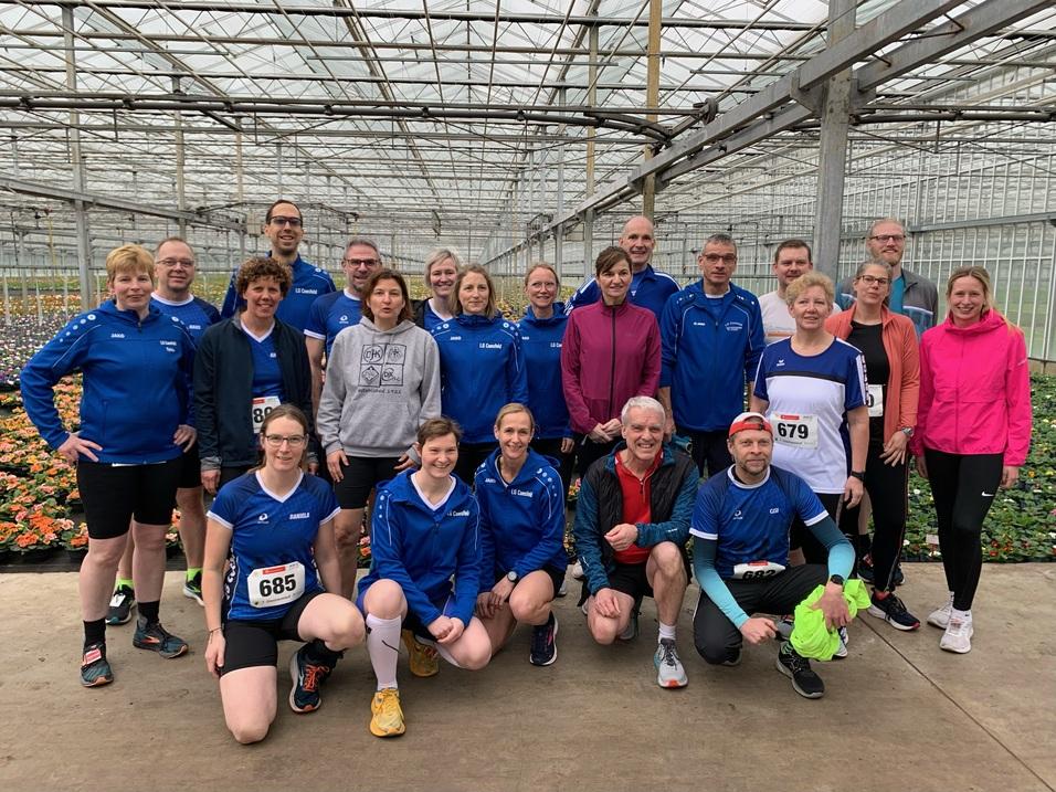 Gruppe von 20 Läufern in blauen Trikots steht in einem Gewächshaus zwischen bunten Blumen.