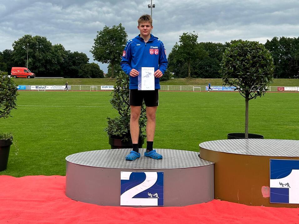 Junge auf dem zweiten Platz steht auf dem Siegertreppchen, hält eine Urkunde, im Hintergrund ein Sportplatz unter bewölktem Himmel.
