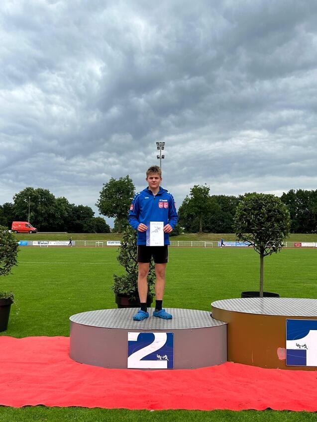 Junge auf dem zweiten Platz steht auf dem Siegertreppchen, hält eine Urkunde, im Hintergrund ein Sportplatz unter bewölktem Himmel.