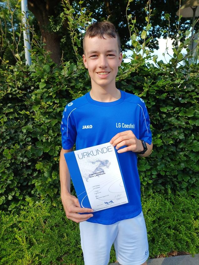 Junger Sportler in blauem Trikot hält stolz eine Urkunde vor einer grünen Hecke.