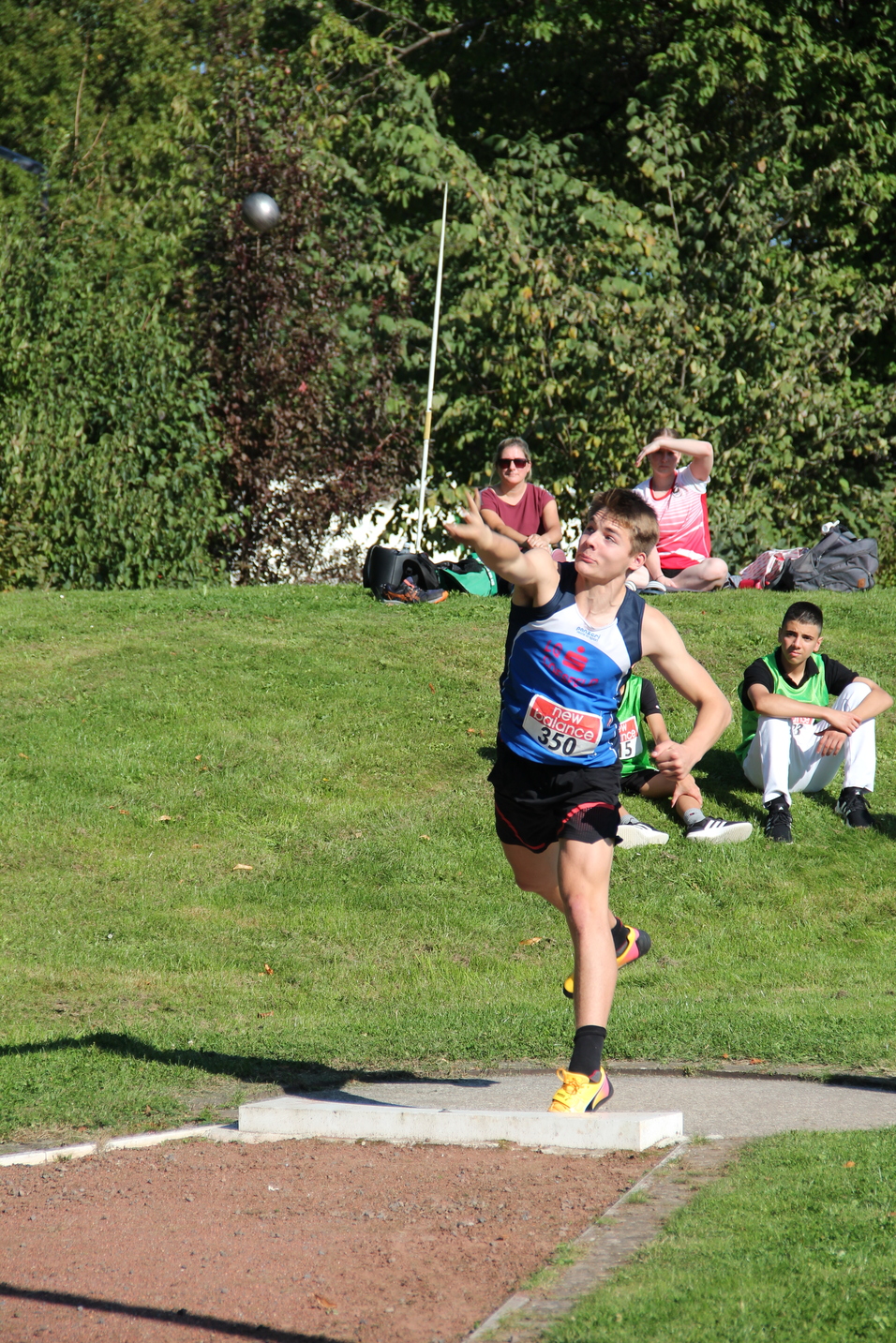Athlet wirft eine Kugel beim Wettkampf, während Zuschauer im Hintergrund auf einer Wiese sitzen.