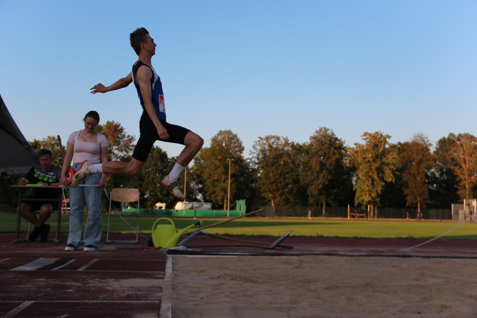 Ein Springer in Sportkleidung vollzieht einen Satz über eine Sandgrube, während zwei Zuschauer aufmerksam beobachten.