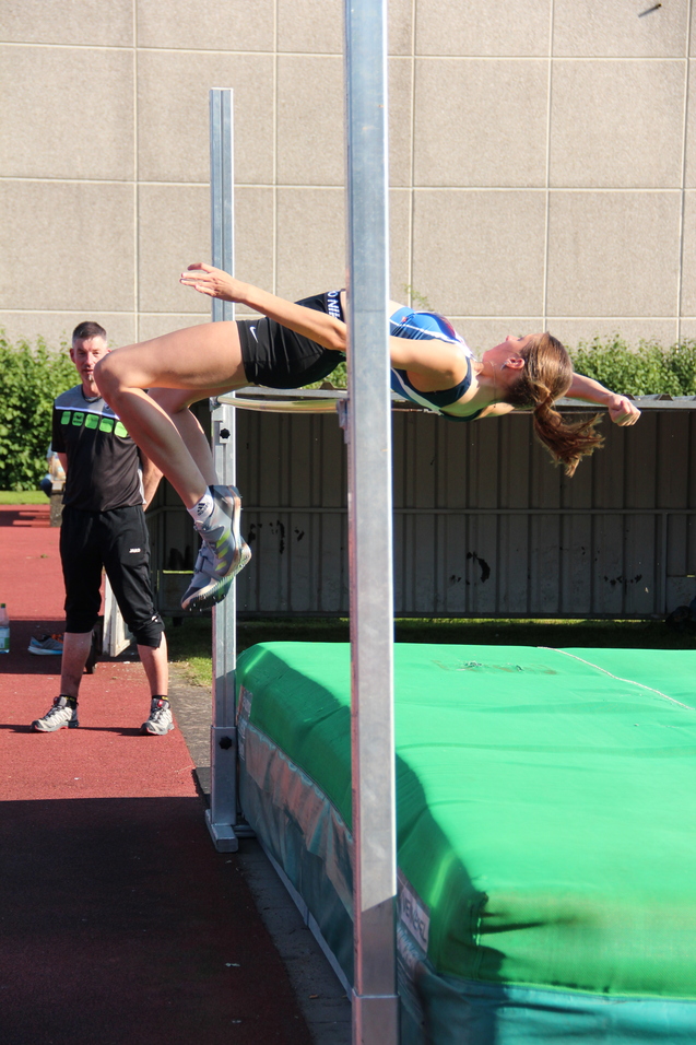 Sportlerin springt Hochsprung über eine Stange, während ein Zuschauer im Hintergrund steht. Wettkampfatmosphäre auf dem Sportplatz.