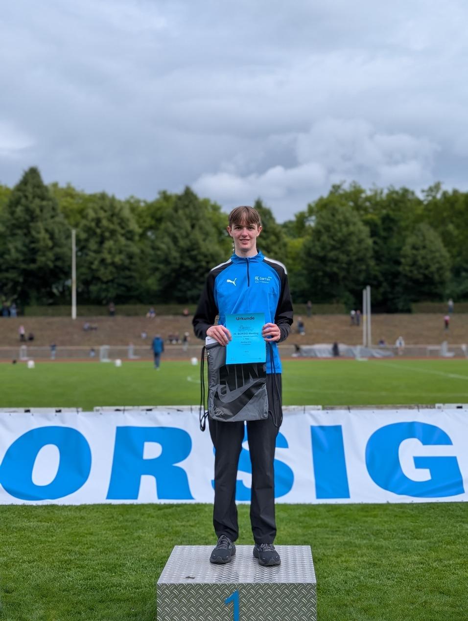 Junger Athlet steht auf dem Siegerpodest, hält eine Urkunde und trägt Sportkleidung in Blau und Schwarz.
