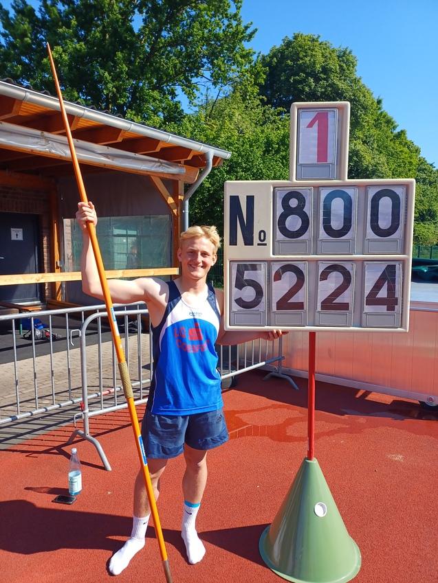 Junger Athlet mit einem Speer steht neben einer Anzeigetafel mit den Zahlen 800 und 5224 auf einer Sportanlage.