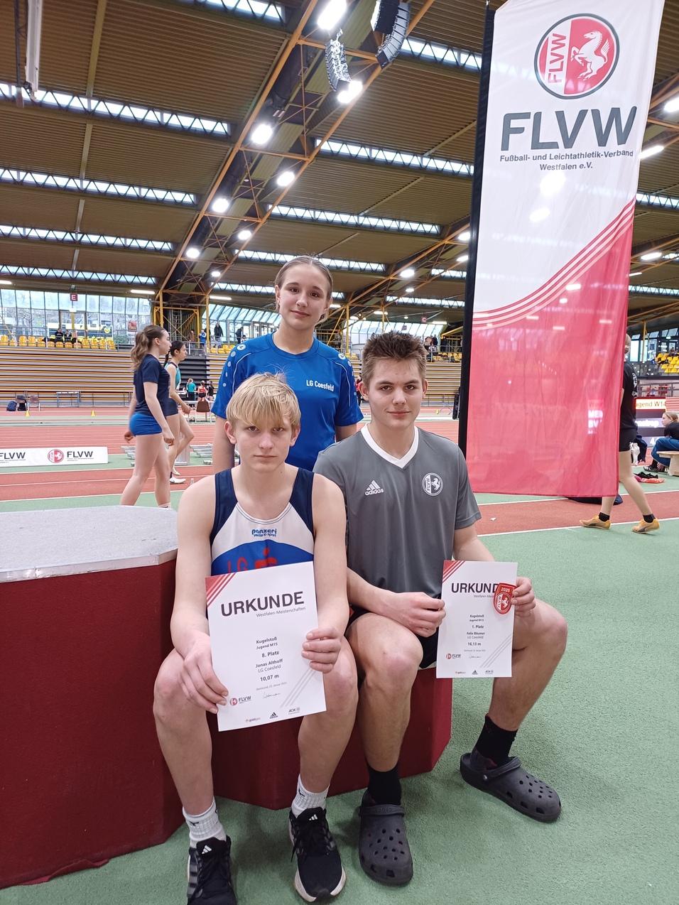 Drei Jugendliche halten Urkunden in einer Sporthalle, umgeben von Sportgeräten und anderen Teilnehmern.