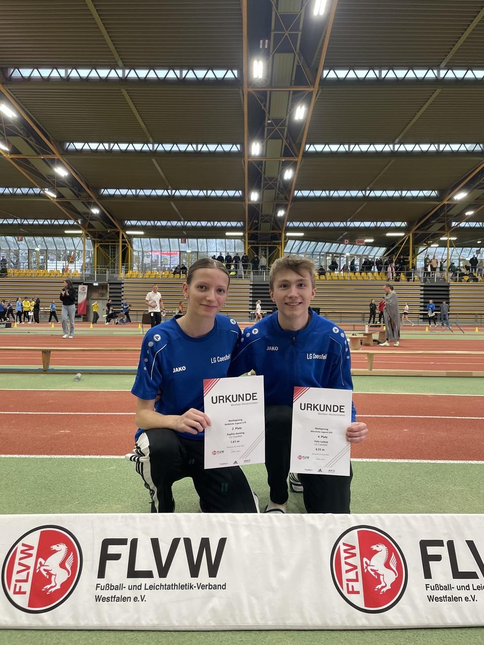 Zwei Athleten in blauen Trikots halten Urkunden in der Sporthalle, mit einer FLVW-Bande im Vordergrund.
