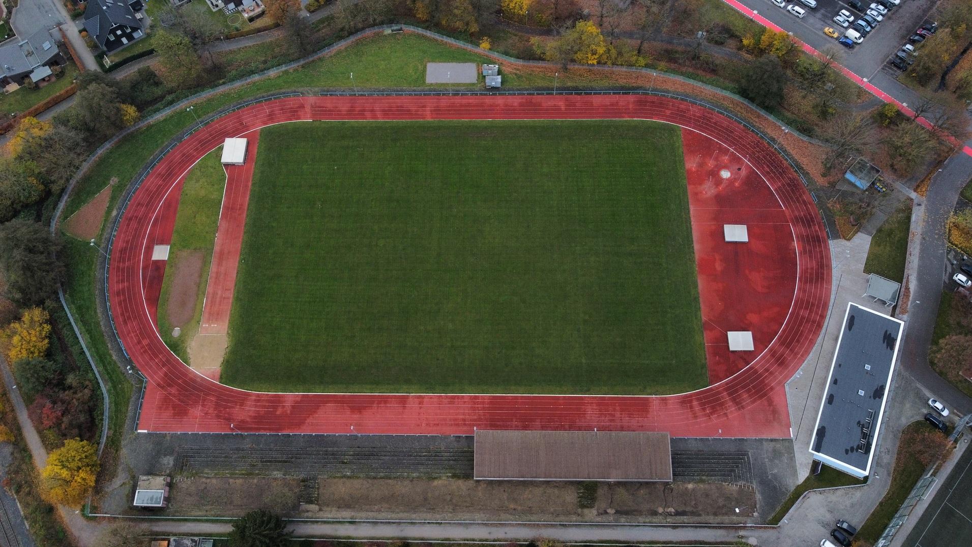 Luftaufnahme eines Sportplatzes mit roter Laufbahn und grüner Rasenfläche, umgeben von Bäumen und Parkplätzen.