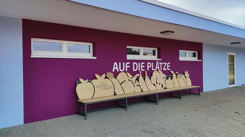 Bunte Wand mit dem Schriftzug „AUF DIE PLÄTZE“ und einer Holzbank mit Obst- und Gemüse-Silhouetten.