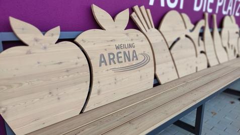 Brett mit Holzgestalten von Obst und Gemüse, beschriftet „WEILING ARENA“, vor violettem Hintergrund.