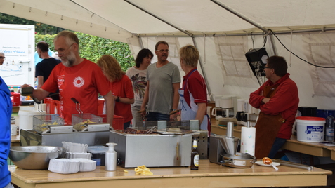 Menschen in einem Festzelt stehen um eine große Arbeitsfläche mit Kochutensilien und Lebensmitteln.