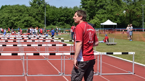 Mann in roter Sportbekleidung steht vor einer Hindernisbahn, mit Wettkämpfen im Hintergrund auf einem Sportgelände.