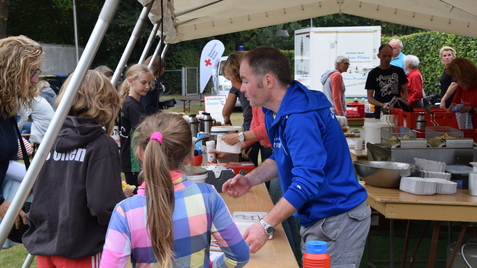 Menschen stehen an einem Tisch unter einem Zelt, um Essen und Getr&auml;nke bei einer Veranstaltung zu erhalten.