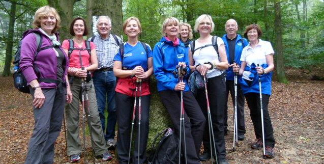 Gruppe von neun Wanderern mit Stöcken in einem Wald, umgeben von grünen Bäumen und herbstlichem Laub.