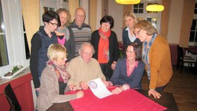 Gruppe von Männern und Frauen steht um einen Tisch, betrachtet ein Dokument auf rotem Tischdecken.