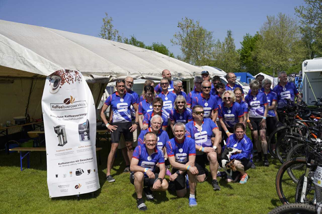 Gruppe von Radfahrern in blauen Trikots, stehend vor einem Zelt mit einem Kaffee-Service-Plakat im Hintergrund.