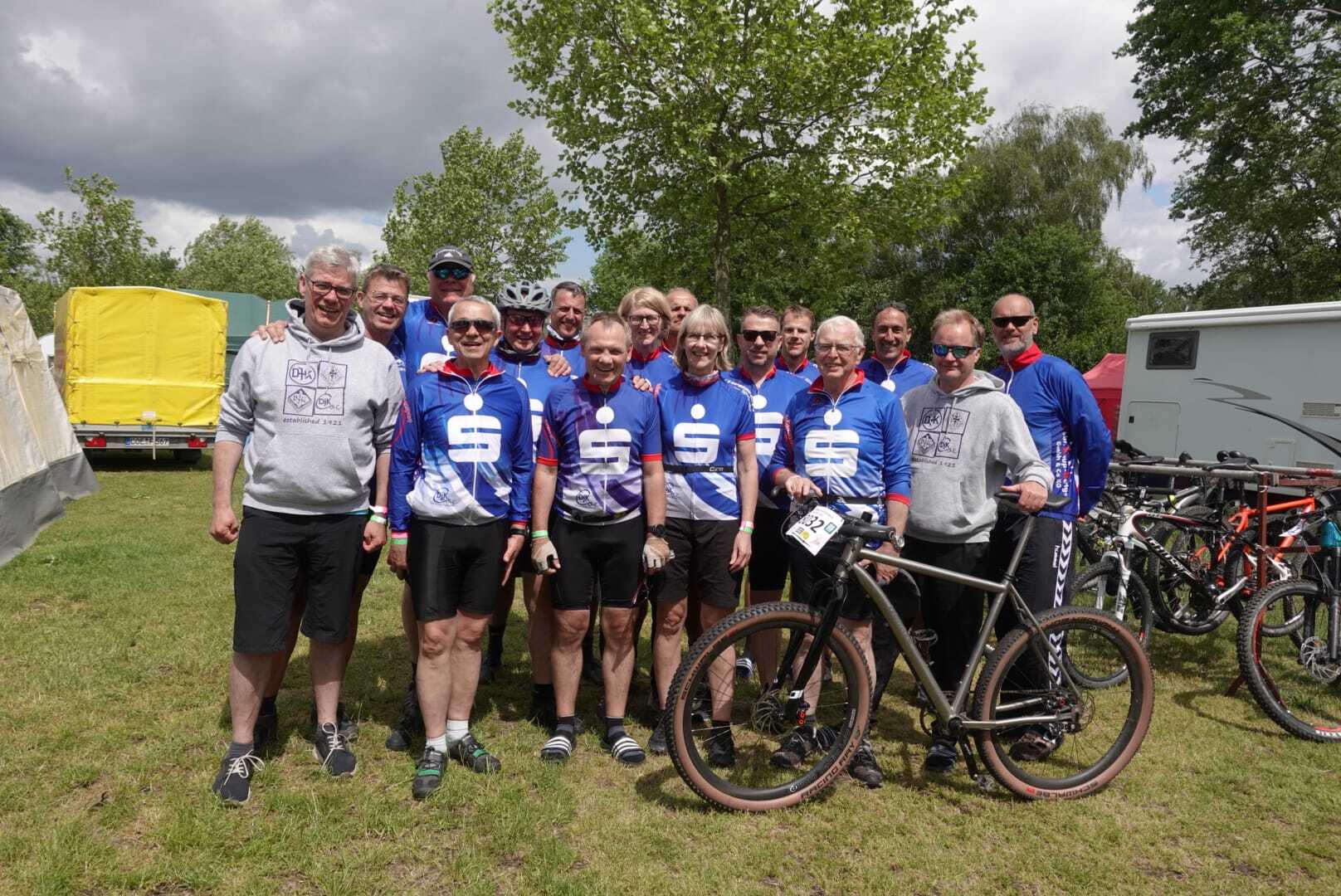 Gruppe von Radfahrern in blauen Trikots posiert mit Mountainbike auf einer Wiese, im Hintergrund Bäume und Wohnwagen.